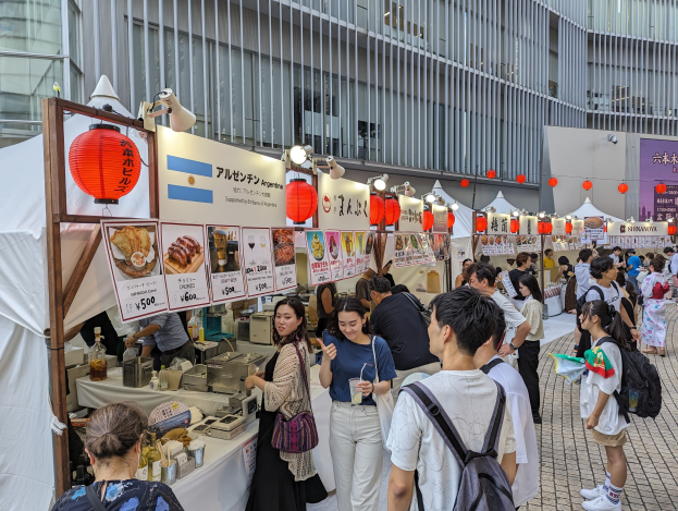 Eine Gruppe von Menschen auf einem Food-Festival in Japan, die um einen Tisch mit Essen versammelt sind, einige tragen Taschen und halten Gläser, während andere die Gegenstände auf dem Tisch untersuchen; Tafeln mit Text und Bildern, Lichter und Laternen hängen von der Decke, mit einem Gebäude im Hintergrund.