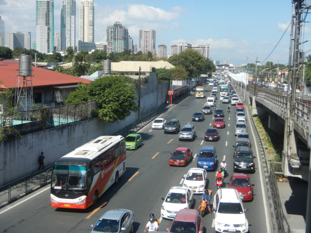 Eine belebte Stadtstraße mit Autos, Bussen und Motorrädern, die eine Brücke mit Strommasten und Drähten, Gebäude, Bäume und einen bewölkten Himmel im Hintergrund zeigt.