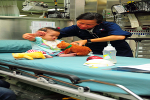 Eine Frau, wahrscheinlich eine Krankenschwester, hölt ein Baby in einem Krankenhauszimmer voller Spielzeuge, einer Flasche, einer Kappe und anderen Gegenständen, mit medizinischen Geräten und einem Vorhang im Hintergrund.