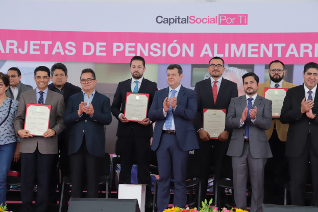 Eine Gruppe von Menschen auf einer Bühne, die klatschen und Zertifikate halten, mit Stühlen in einem Halbkreis drumherum und einem Banner mit der Aufschrift "Capital Social for Ti" im Hintergrund, sowie Blumensträuße und Redner am Boden.