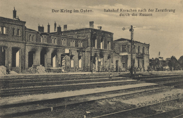Ein altes Schwarz-Weiß-Foto eines Bahnhofs in Korschen, Deutschland, das Schienen, Gebäude, Pfosten, Menschen, Bäume und einen Himmel im Hintergrund zeigt, mit dem Text "Bahnhof Korschen in Ostern, Deutschland" oben und unten auf dem Bild.