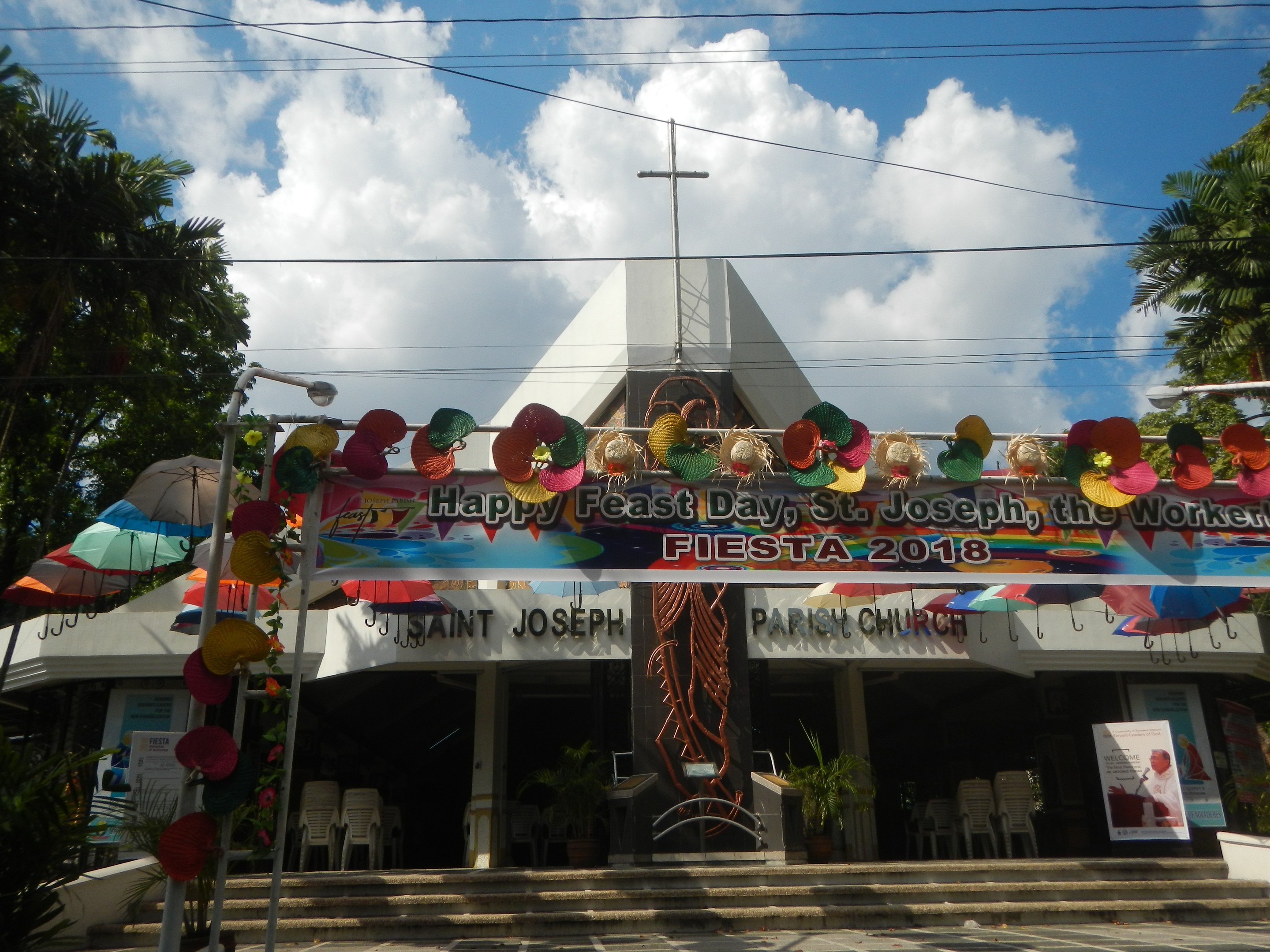 Ein Gebäude mit einem Schild "Frohes Fest St. Josef der Arbeiter" davor, mit einer Treppe, die zu einem Eingang mit einem gewölbten Schmuckrahmen führt, Außenbestuhlung, Topfpflanzen, Bäumen und einem bewölkten Himmel.