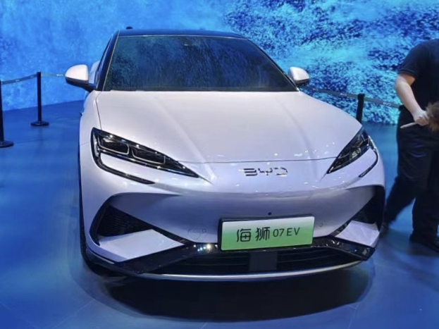 Ein weißes Auto auf einer Automesse mit einer Person rechts daneben, die ein Objekt hält, einem Bildschirm im Hintergrund und Absperrpollern mit Seilen links.