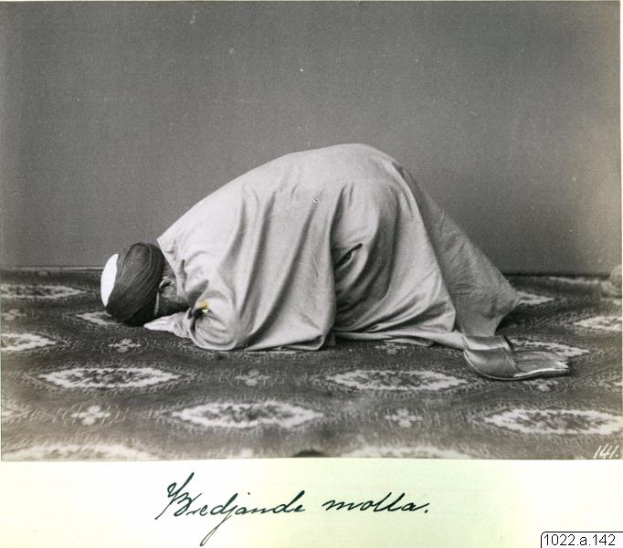 Ein altes Schwarz-Weiß-Foto eines Mannes, der betend kniet und ein Tuch und eine Kappe trägt, mit der Aufschrift "Bredjande Molla, die erste muslimische Frau der Welt" unten.