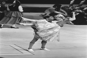 Ein Schwarz-Weiß-Foto eines Paares, das auf einer Bühne tanzt, mit ein paar Menschen auf einer Treppe im Hintergrund und einem Pfahl im Vordergrund.