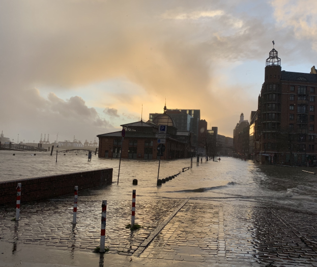 Überschwemmte Straße in Hamburg, Deutschland, mit Wasser auf der Straße, Polen, Schildern, Gebäuden, einer Brücke und einem bewölkten Himmel im Hintergrund.