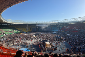 Eine große Menschenmenge versammelt sich in einem Stadion, mit einer Bühne in der Mitte, auf der verschiedene Gegenstände zu sehen sind, und einem Dach mit Lichtern oben, was auf ein Konzert im Wembley-Stadion hindeutet.