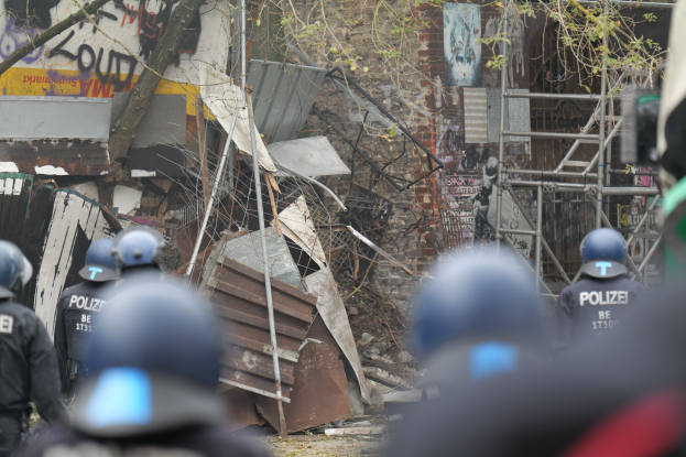 Eine Gruppe von Polizisten in Helmen steht vor einem teilweise abgerissenen Gebäude mit Text an seiner Wand, umgeben von Bäumen und Pfählen.