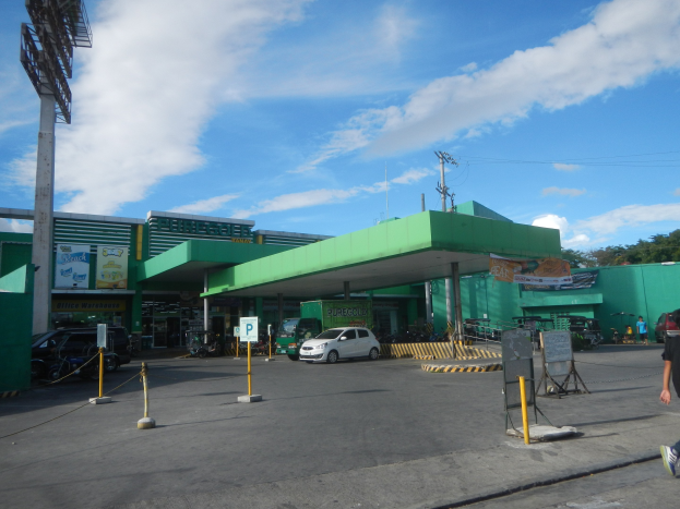 Ein Mann geht an einer grünen Tankstelle vorbei mit Fahrzeugen, Absperrpollern, Schildern, Werbetafeln, Strommasten, Bäumen und einem bewölkten Himmel im Hintergrund.
