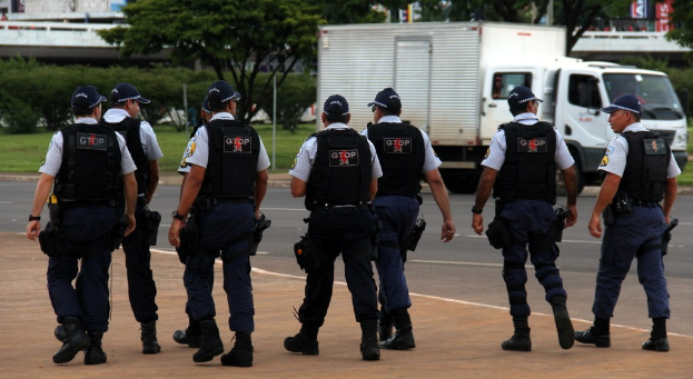 Eine Gruppe von Beamten in Uniformen, darunter Mötzen, Hemden, Schutzwesten, Uhren, Hosen und Schuhe, die auf einer Straße mit einem geparkten Lastwagen rechts, Gebäuden, Bäumen, Pflanzen, Masten, Bannern und einer Brücke im Hintergrund.
