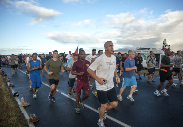 Gruppe von Läufern bei einem Marathon auf einer Straße mit Gras auf beiden Seiten, die sportliche Schuhe tragen, mit Fahrzeugen, Bäumen, Hügeln und einem bewölkten Himmel im Hintergrund.