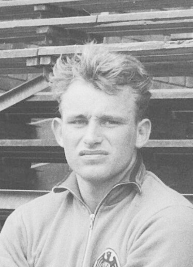 Schwarzes und weiße Foto eines Mannes in einer Jacke, der vor einem Stapel hölzerner Bretter steht, der als legendärer deutscher Füssballer gilt.