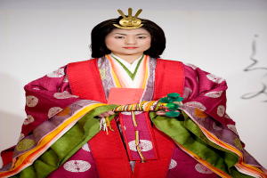 Eine Frau in einem traditionellen japanischen Kimono, das ein leuchtend rotes und grünes Kleid mit kunstvollen Mustern trägt, steht selbstbewusst auf einem Laufsteg, ihr Haar zu einem ordentlichen Knoten gebunden.