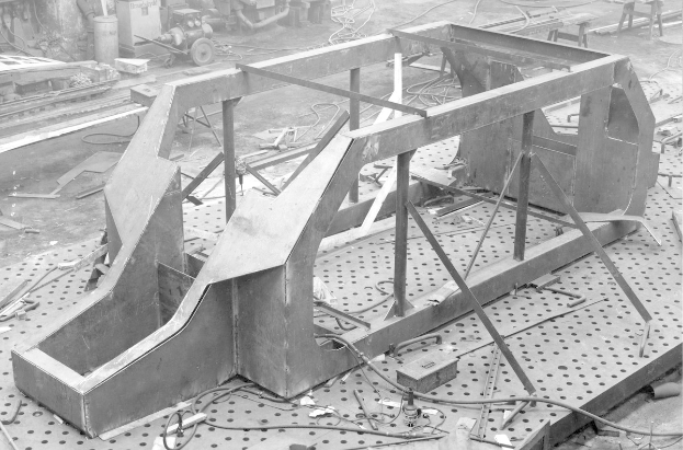 Ein Schwarz-Weiß-Foto eines Autoschassis auf einem Fabrikboden, umgeben von verstreuten Werkzeugen und Maschinen im Hintergrund.