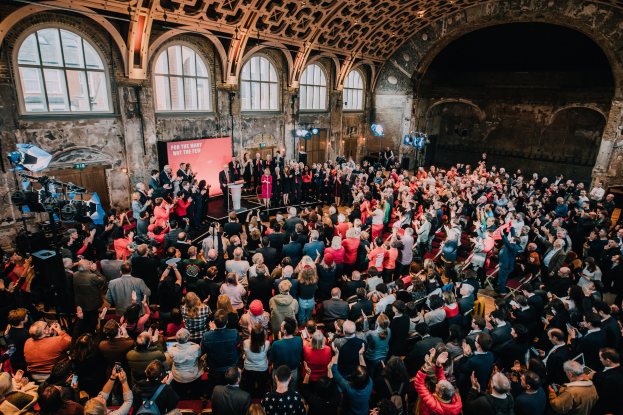 Große Menschenmenge in einem Gebäude mit gewölbten Fenstern und einer Bühne, auf der einige Menschen stehen; Kameras und Equipment sind links zu sehen, und eine Wand im Hintergrund zeigt "The Great Gatsby Awards 2019" mit einer sichtbaren Decke darüber.