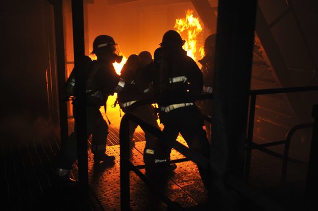 Feuerwehrleute in Schutzausrüstung vor einem brennenden Gebäude mit Flammen und Rauch am Nachthimmel