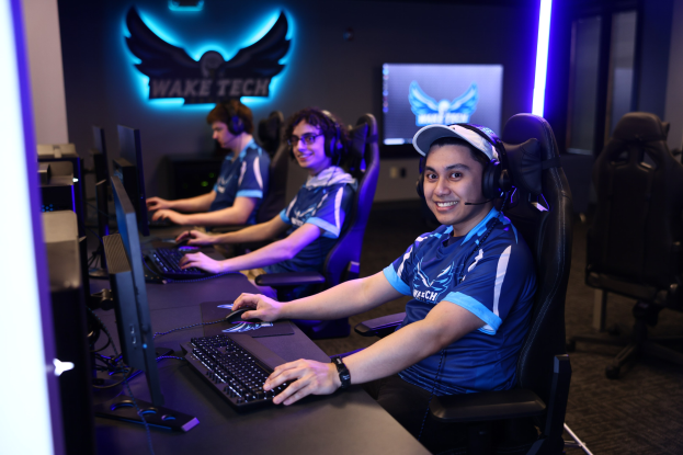 Eine Gruppe von Menschen mit Kopfhörern sitzt an einem Schreibtisch mit Computern, Monitoren, Tastaturen und Mäusen, die vor einer Wand mit Logo und Text lächeln, wobei Stühle rechts zu sehen sind.