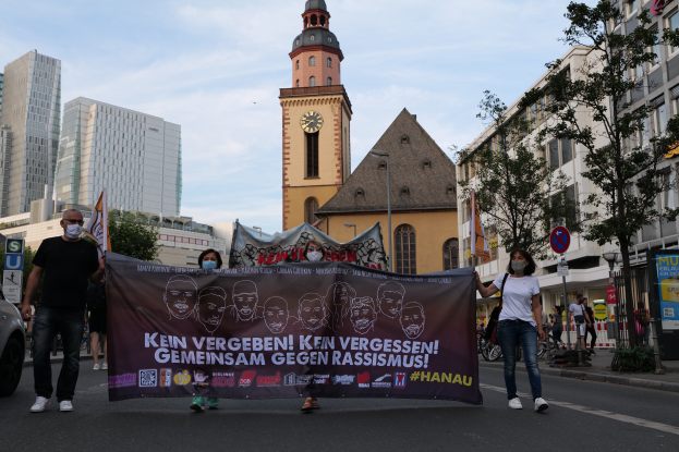 Eine Gruppe maskierter Menschen marschiert die Straße entlang und hölt eine Protestfahne, mit einem geparkten Auto auf der linken Seite, Gebäude, Bäume, Schilder, Masten und ein Uhrenturm im Hintergrund unter einem klaren blauen Himmel.