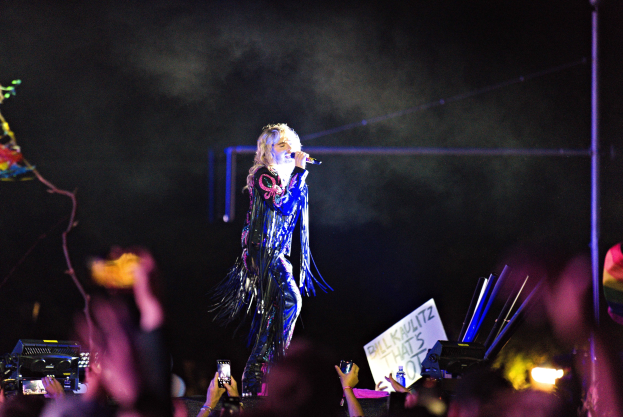 Eine Frau auf einer Bühne hält ein Mikrofon, während eine Menge darunter sie mit Handys aufnimmt oder bewundernd nach oben schaut, vor einem dunklen Hintergrund.