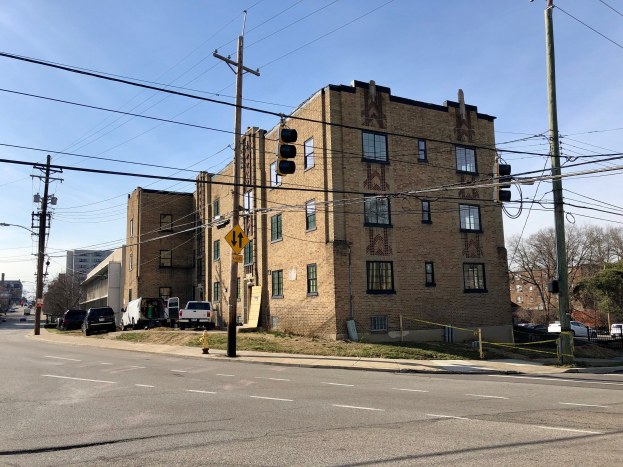 Großer Backsteinwohnblock an einer Straßenkreuzung mit Fahrzeugen, Ampeln, Schildern, Strommasten und Bäumen im Hintergrund.
