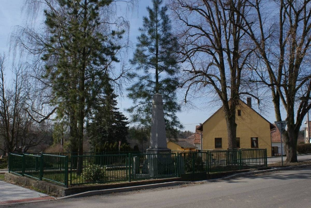 Denkmal für Holocaust-Opfer in der Mitte einer Straße, umgeben von einem Metallzaun, Grünfläche, Häusern, einem Straßenpfahl und einem bewölkten Himmel.