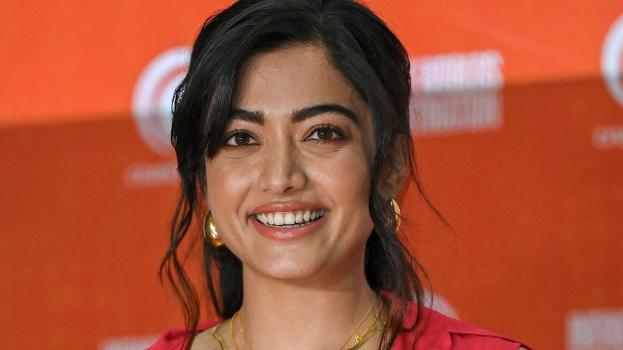 Eine Frau in einem roten Kleid mit einem eleganten Dutt und dezentem Make-up lächelt in die Kamera, trägt schwarze High Heels und hat einen hellgelben Banner mit der Aufschrift 'Die besten Schönheits Looks von Sonam Kapoor beim Cannes Film Festival' im Hintergrund.
