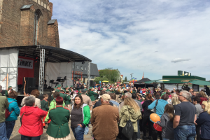 Eine große Menschenmenge steht vor einer Kirche, einige halten Luftballons, mit einem Gebäude auf der linken Seite, Musikinstrumenten und einem Banner auf einer Bühne und Bäumen und einem bewölkten Himmel im Hintergrund, was auf ein St. Patrick's Day-Fest hinweist.