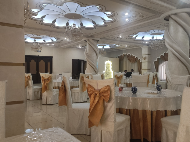 Ein großer Festsaal mit Tischen und Stühlen, die mit Tüchern drapiert und mit verschiedenen Gegenständen geschmückt sind, mit Türen und Vorhängen an den Wänden und einer Decke mit Lampen und Kronleuchtern.