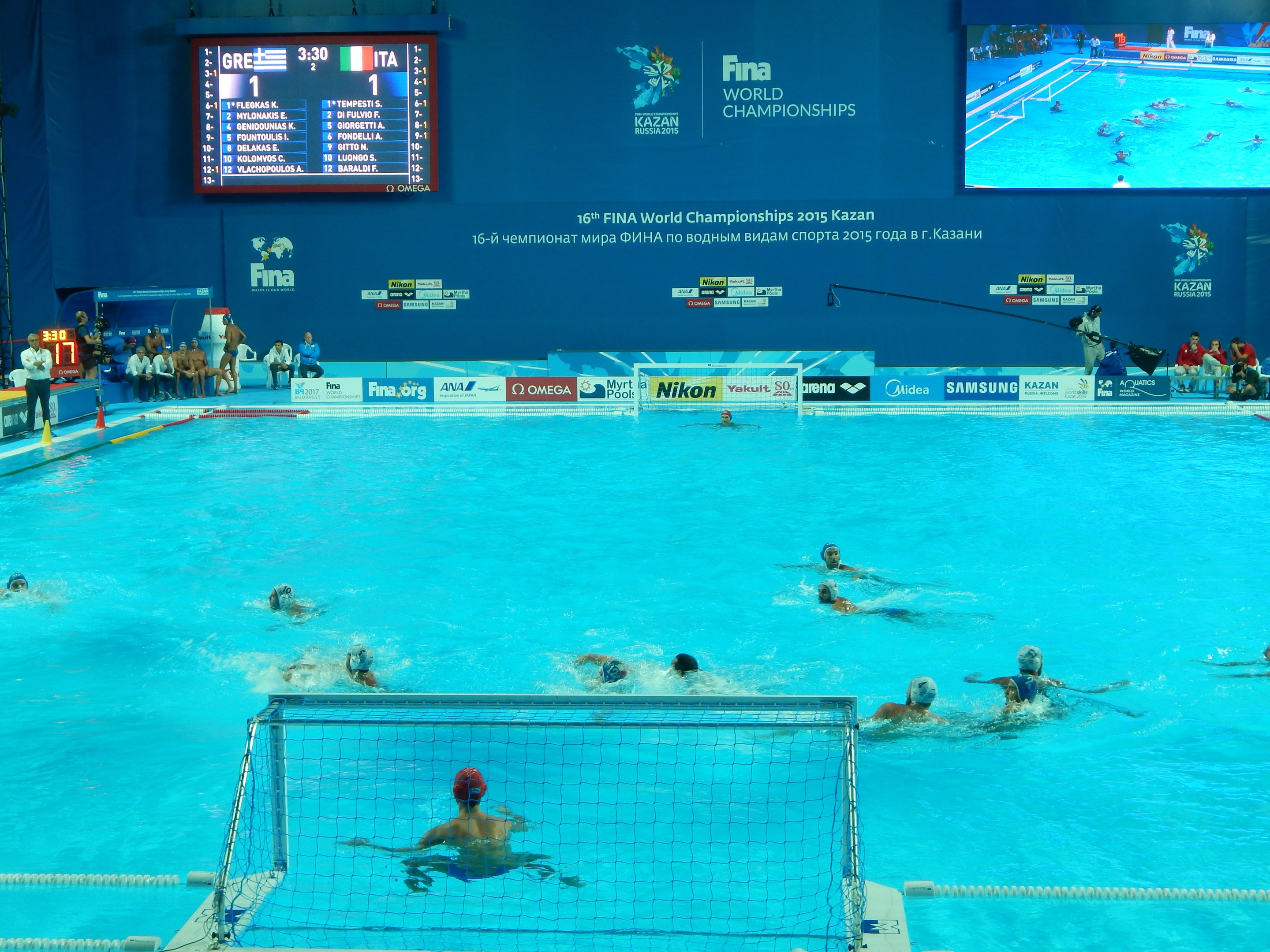 Wasserballspieler bei den 2016 FINA-Weltmeisterschaften in Kazan im Pool gegeneinander antretend, mit Zuschauern auf der linken Seite und einem Scoreboard im Hintergrund.