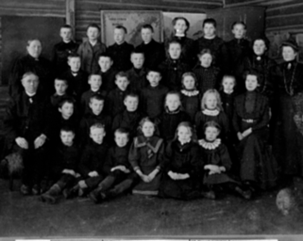 Ein Schwarz-Weiß-Foto einer Gruppe von Kindern, die vor einer Wand mit einer Tafel im Hintergrund posieren, mit dem Text "Die erste Klasse der 1930er Jahre" unten.