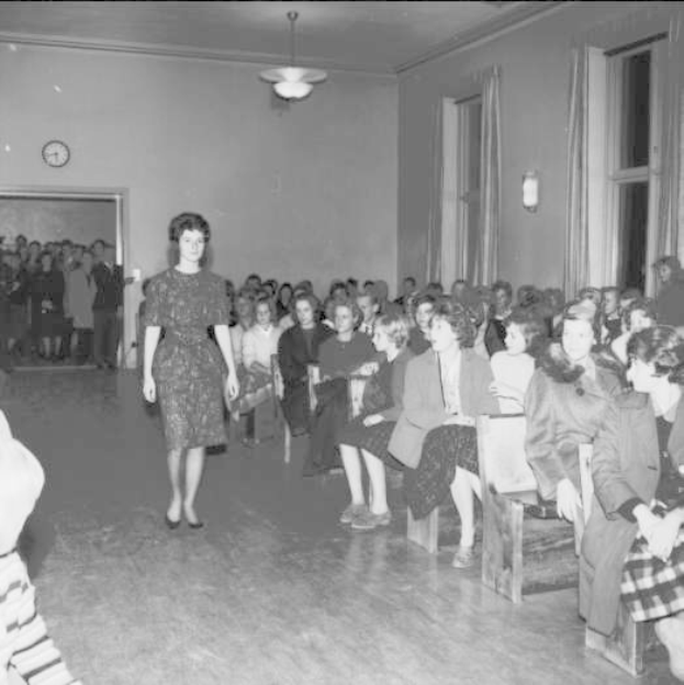 Eine Frau in einem schwarzen und weißen Kleid läuft über einen Laufsteg während einer Modenschau für Frauenrechte, mit einer Menge, die sitzt und zusieht, einer Uhr und Lampen an der Wand und einer beleuchteten Decke.