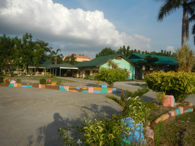Ein Resort mit zahlreichen Palmen im Vordergrund, umgeben von Häusern, Pflanzen und Gras, mit einer Straße in der Nähe und einem Himmel voller weißer, flauschiger Wolken im Hintergrund.