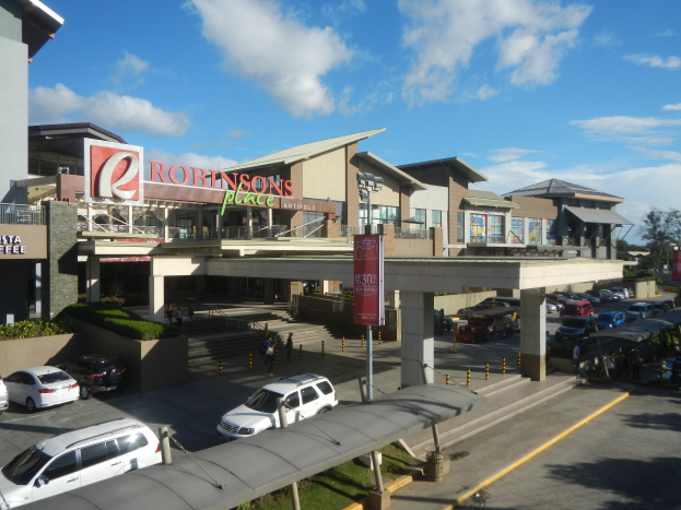 Ein belebter Einkaufszentrum mit verschiedenen Gebäuden, Geschäften, Fahrzeugen auf der Straße, Fußgängern auf dem Gehweg, Straßenlaternen, Informationen, Grünflächen und einem bewölkten Himmel.