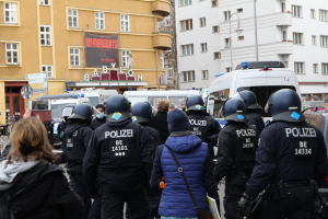 Eine Gruppe von Polizeibeamten steht vor einer Menschenmenge, die Helme und Jacken trägt, während einer Demonstration in Berlin, Deutschland, mit Fahrzeugen, Gebäuden, Laternenpfählen und einem Banner mit Text im Hintergrund und einer Person mit einer Kamera.