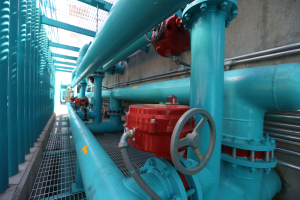 Eine große Gruppe von blauen Rohren und Ventilen in einem Gebäude, mit einem prominenten roten Ventil in der Mitte, Metallstangen auf der linken Seite und einer Wand im Hintergrund.