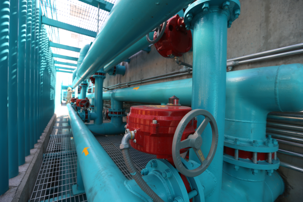 Eine große Gruppe von blauen Rohren und Ventilen in einem Gebäude, mit einem prominenten roten Ventil in der Mitte, Metallstangen auf der linken Seite und einer Wand im Hintergrund.