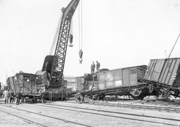 Schwarzes und weißes Bild einer Zugkatastrophe mit einem Kran, der ein Zugwaggon auf die Schienen hebt, mehrere Menschen in der Nähe und Gebäude im Hintergrund bei klarem Himmel.