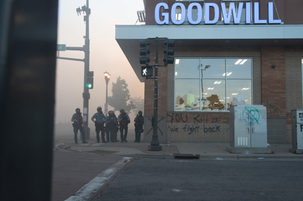 Eine Gruppe von Polizisten in Helmen und Ausrüstung steht mit Waffen vor einem Second-Hand-Laden mit Glaswänden und Beschriftung, während Laternenmasten, Ampeln, Bäume und Himmel im Bild zu sehen sind.
