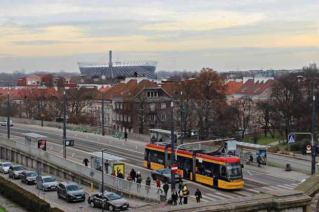 Eine gelbe und schwarze Straßenbahn fährt auf einer Stadtstraße mit hohen Gebäuden, umgeben von Autos, Fußgängern, Bäumen und Straßeninfrastruktur unter einem bewölkten Himmel.