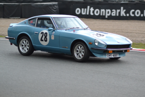 Ein blauer Wagen fährt auf einer Rennstrecke mit einem Helmträger am Steuer, umgeben von Reifen, einer Fahne und Gras.
