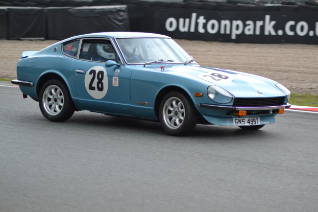 Ein blauer Wagen fährt auf einer Rennstrecke mit einem Helmträger am Steuer, umgeben von Reifen, einer Fahne und Gras.