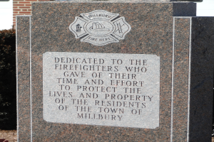 Denkmalstein mit einer Gedenktafel für Feuerwehrleute in Millbury, vor einem Backsteingebäude mit umliegender Vegetation.