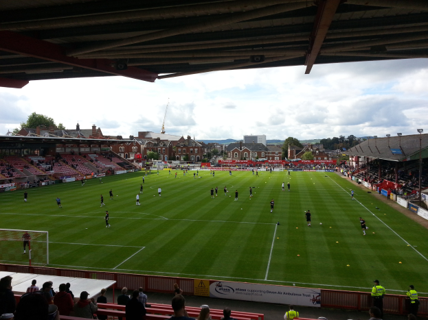 Ein Fußballspiel im St. Mary's Stadium mit Zuschauern auf den Tribünen und Stehplätzen, ein Torpfosten auf der linken Seite und Gebäude, Bäume und einen bewölkten Himmel im Hintergrund.