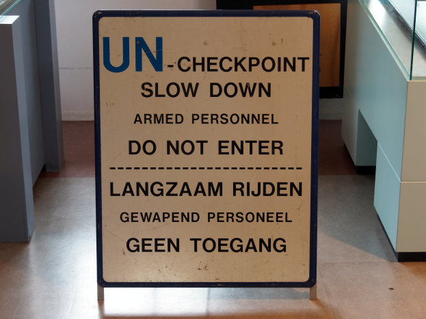 Schild auf dem Boden mit der Aufschrift "UN-Checkpoint Verlangsame bewaffnete Person - Betreten verboten" mit einer Glasplatte und Gegenständen rechts und einer Wand mit einem Gegenstand im Hintergrund.