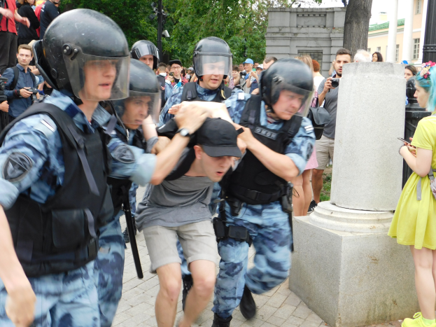 Eine Gruppe von Bereitschaftspolizisten in Ausrüstung, die vor einer Menge davonläuft, die Helme, Waffen und Kameras hält, mit einer Säule im Vordergrund und Gebäuden, Bäumen und Laternen im Hintergrund.