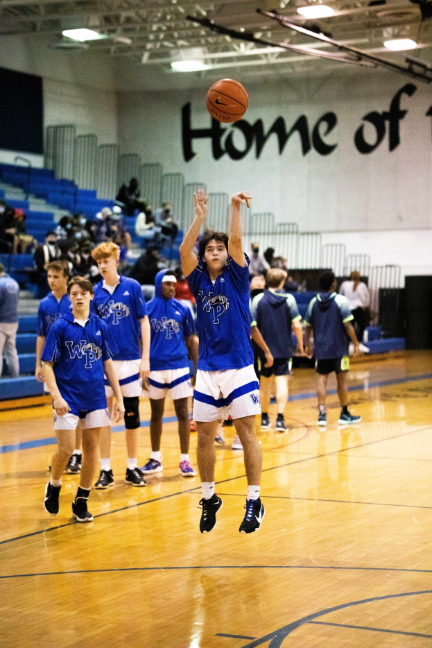 Junge Männer spielen Basketball auf einem Außenplatz mit einer Wand, Treppen, Geländern, Deckenleuchten und Zuschauern, die Turnschuhe tragen, im Hintergrund.